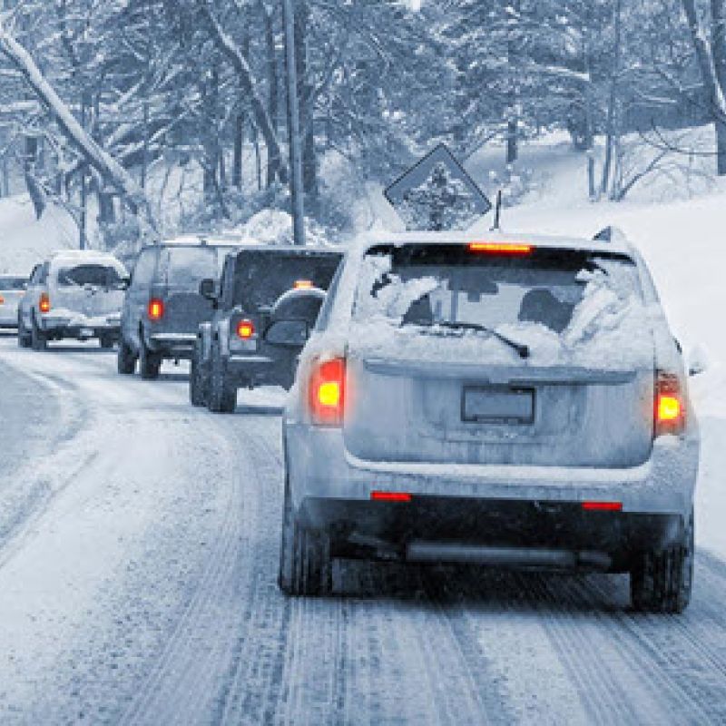 Traffic on snowy road in winter, cars moving slowly due to hazardous conditions, snow-covered trees in the background.