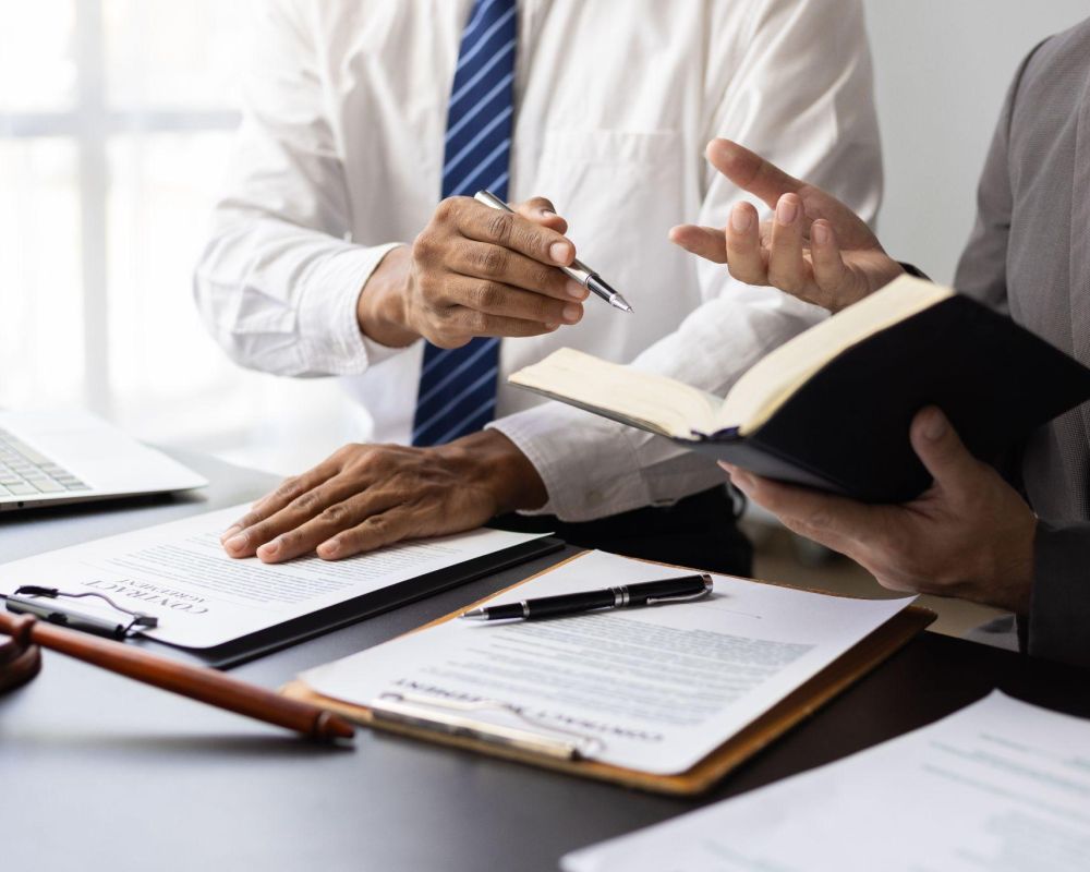 Two professionals discussing legal documents at a desk, with laptop and gavel visible.