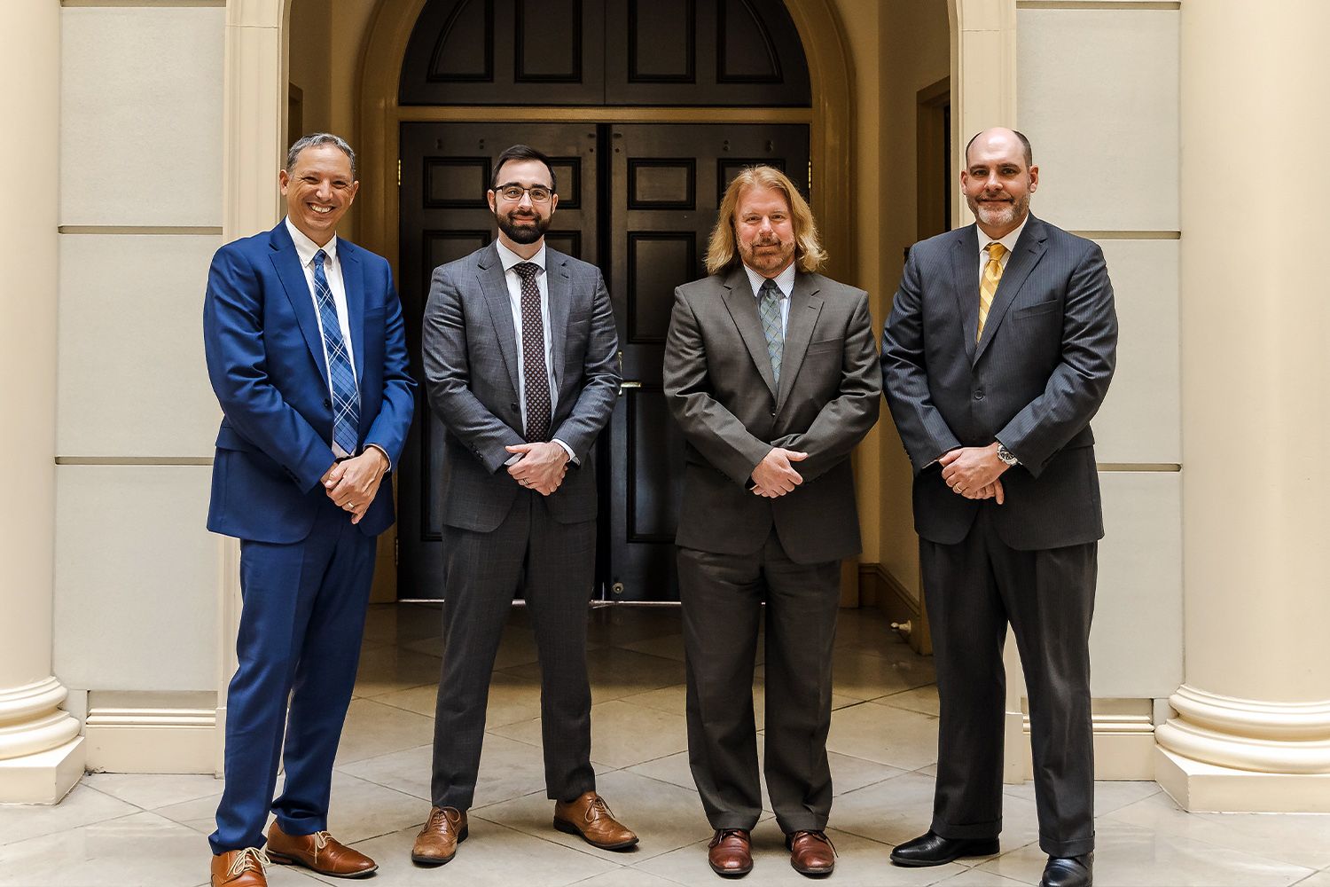 Four businessmen in suits stand together in a hallway, smiling confidently.