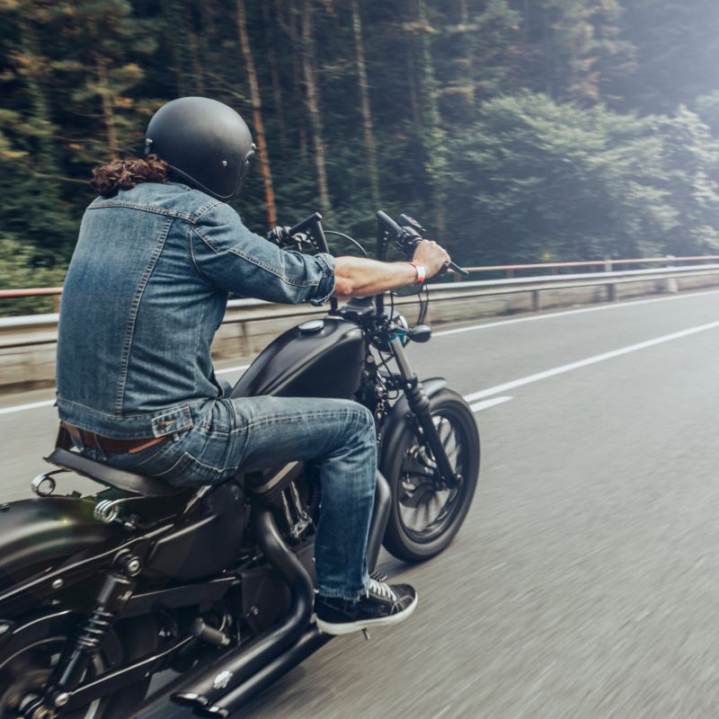 Motorcycle rider wearing a helmet, jean jacket and jeans, riding on a road near trees