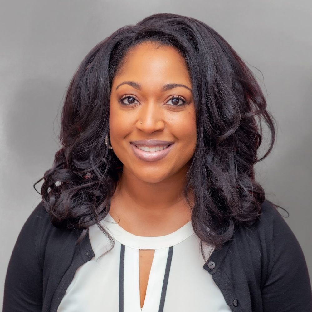 Willonda Ashton smiling in professional attire against a gray background, with long, wavy hair.