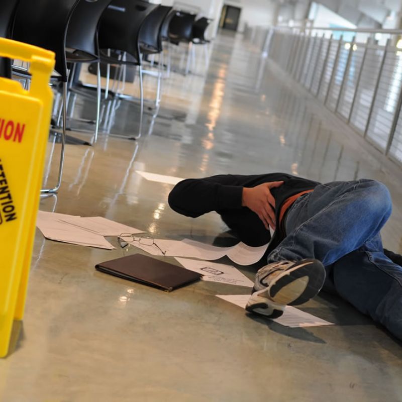 Person slipped on wet floor near caution sign in hallway, surrounded by scattered papers and belongings.