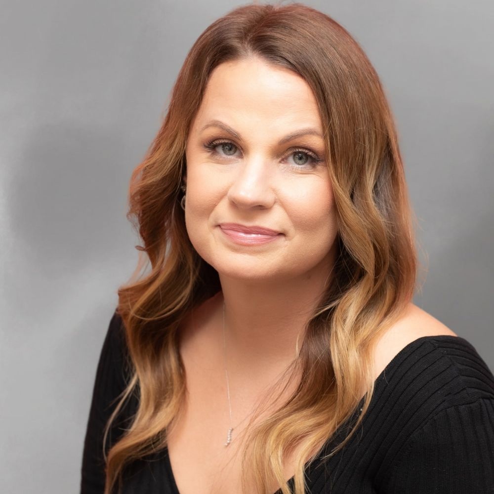 Portrait of Sarah Gray with long hair and a black top, posing against a gray background.