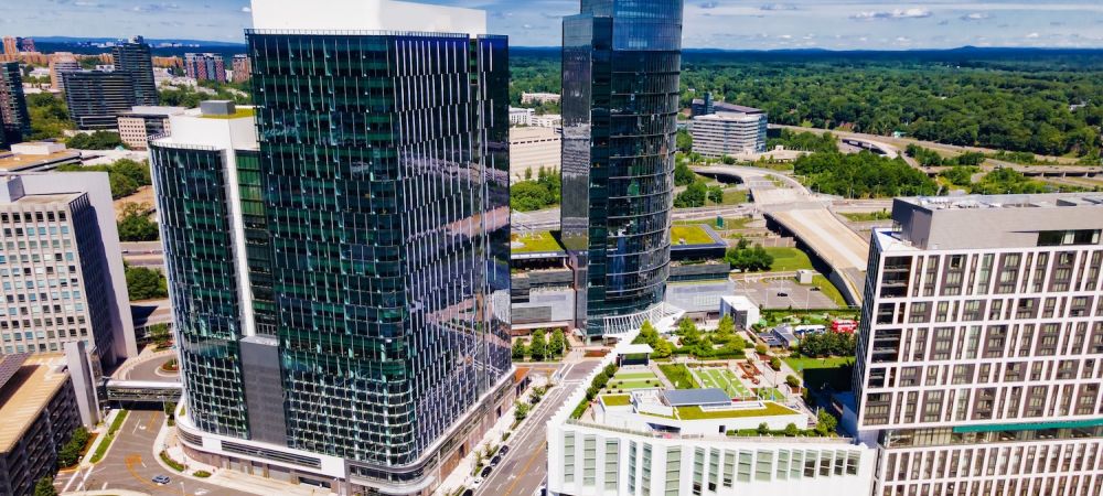 Aerial view of modern glass skyscrapers and lush greenery in an urban landscape under a clear blue sky.