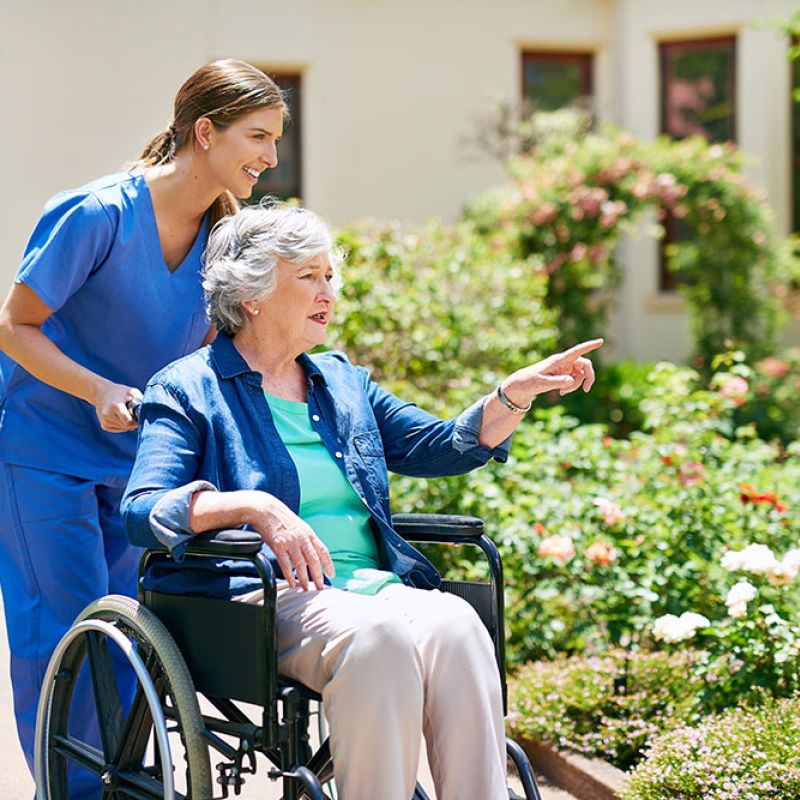Caregiver assisting elderly woman in wheelchair through a garden, both smiling, surrounded by flowers and greenery.