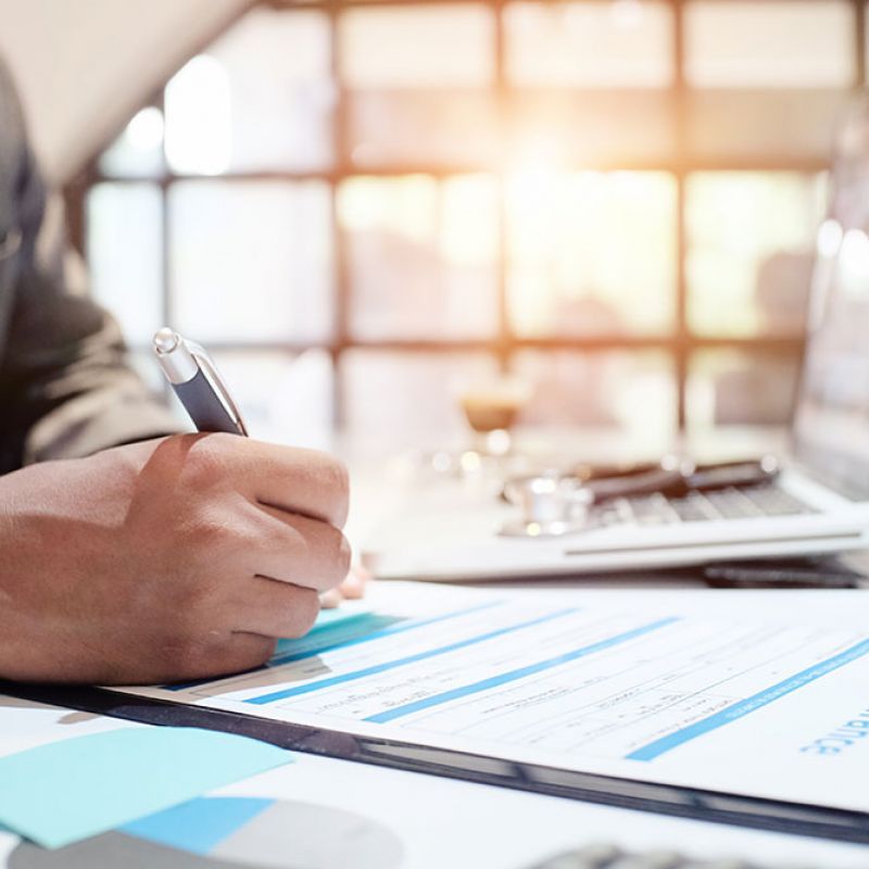Person signing an insurance form at desk with laptop and clipboard, sunlight streaming through window.