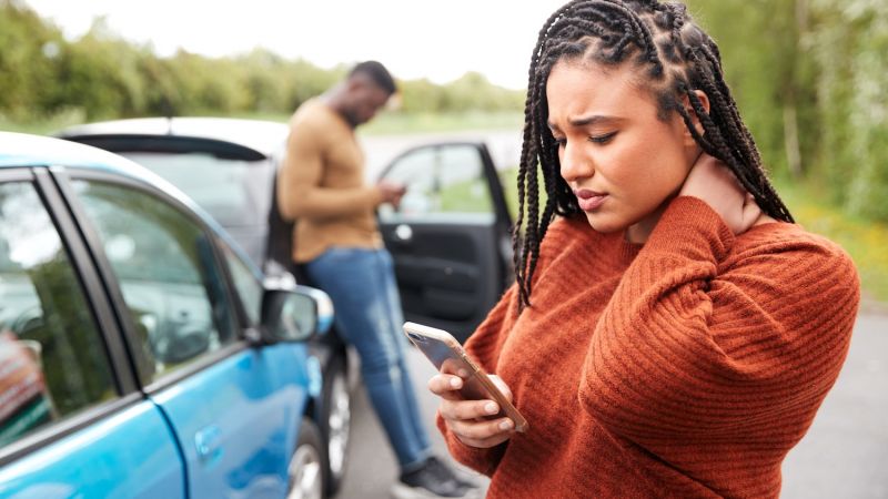 Woman on phone after car collision, rubbing her neck; man in background checking phone near car door.