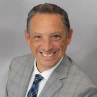 Steve A. Baker in a suit with shoulder-length hair and a patterned tie, smiling in a portrait