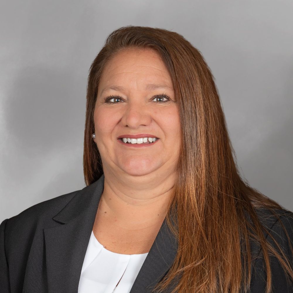 Portrait of Patti Burford with long brown hair in a black blazer smiling against a gray background.