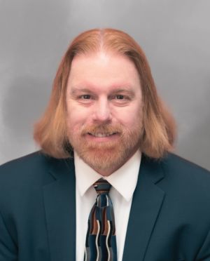 Jonathan DeLoatche with long hair and beard in suit and tie against gray background, smiling confidently.