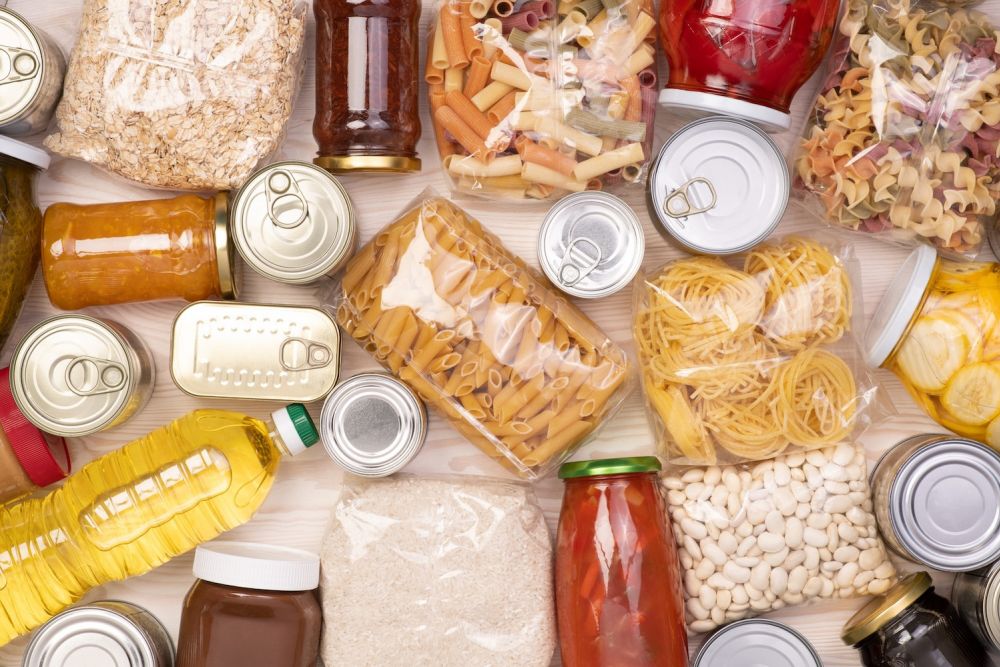 Assorted pantry staples including pasta, canned goods, and bottled items arranged on a wooden surface.
