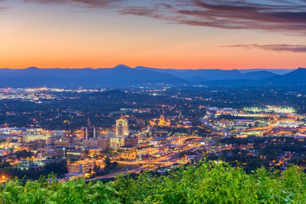 Cityscape of Roanoke, Virginia at sunset, with mountains in the distance and a vibrant skyline illuminated by city lights.