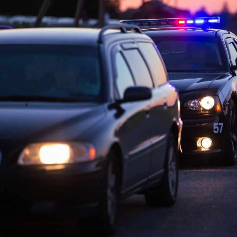 A police car with flashing lights is stopped on the road beside a black vehicle, suggesting a traffic stop during twilight, with fields visible in the background.