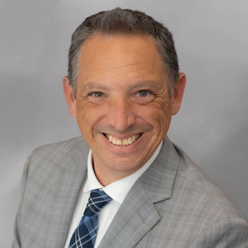 Steve A. Baker in a suit with shoulder-length hair and a patterned tie, smiling in a portrait