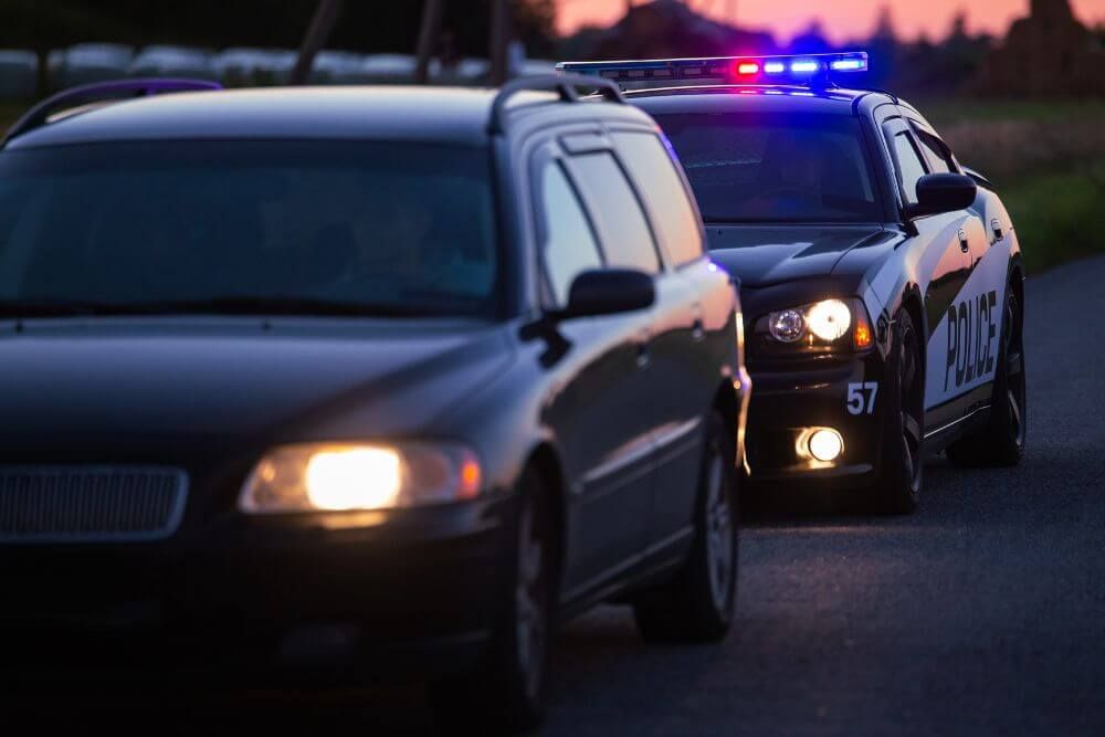 A police car with flashing lights is stopped on the road beside a black vehicle, suggesting a traffic stop during twilight, with fields visible in the background.