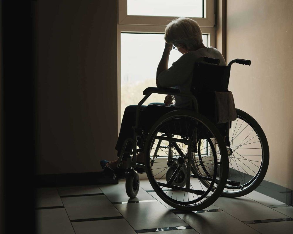 Elderly person in a wheelchair looks out the window, silhouetted in a dim hallway.