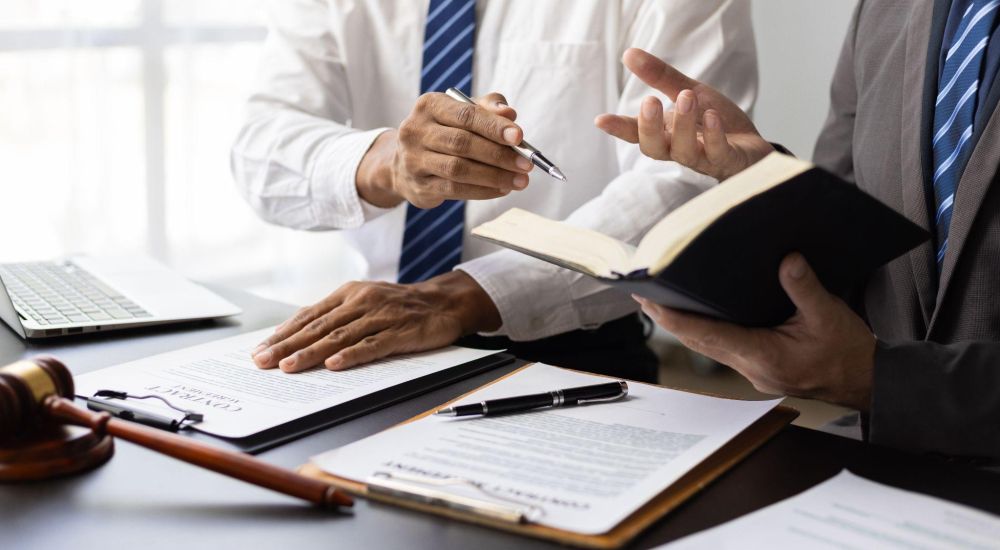 Two professionals discussing legal documents at a desk, with laptop and gavel visible.