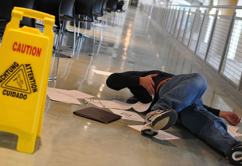Person slipped on wet floor near caution sign in hallway, surrounded by scattered papers and belongings.