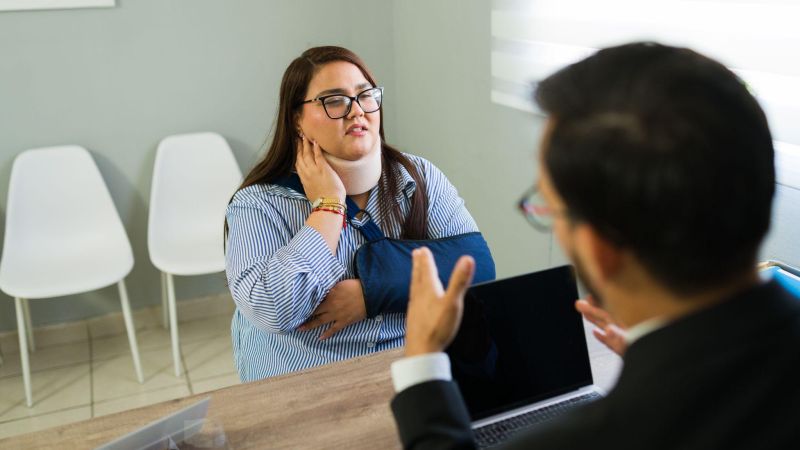 Woman with neck brace and arm sling consults a lawyer in an office setting.