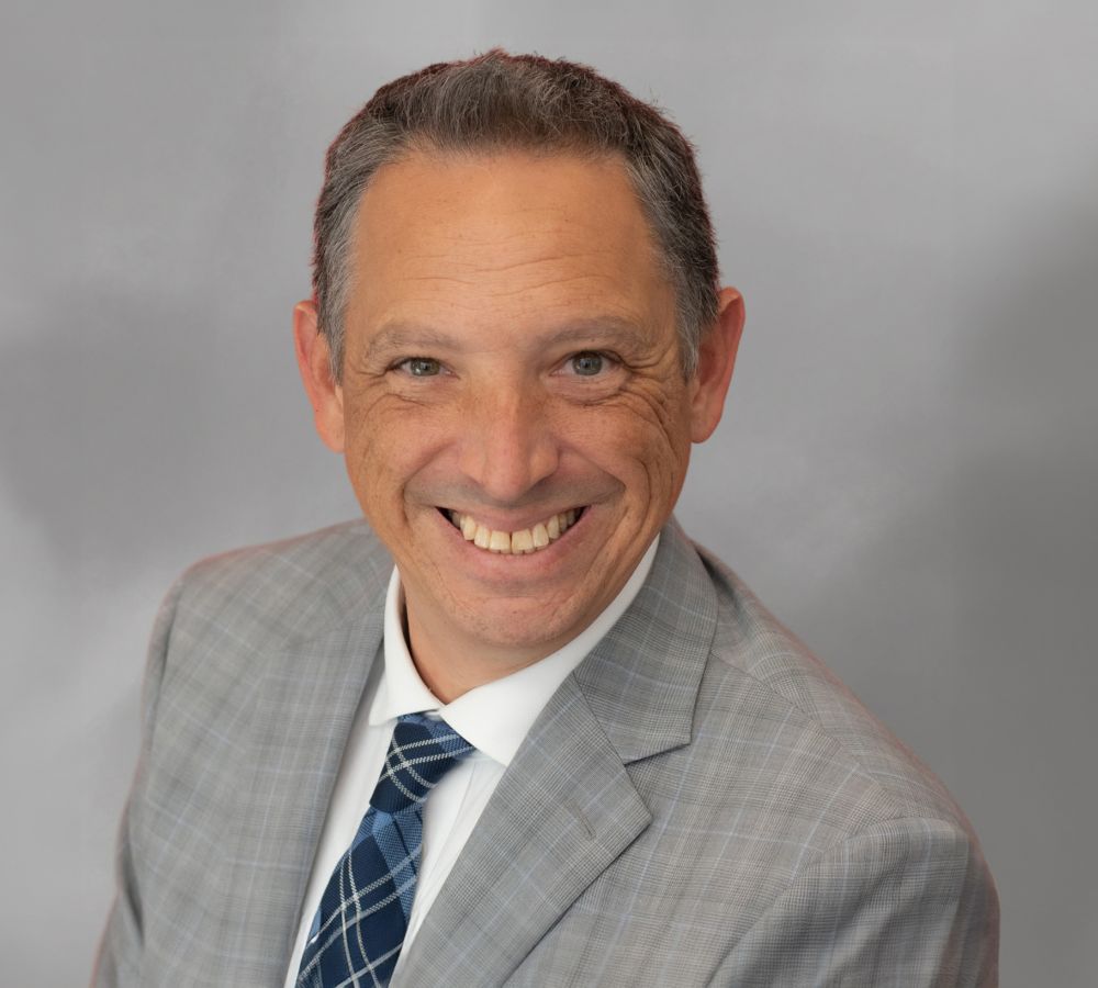 Smiling man in a gray suit with blue striped tie against a gray background.