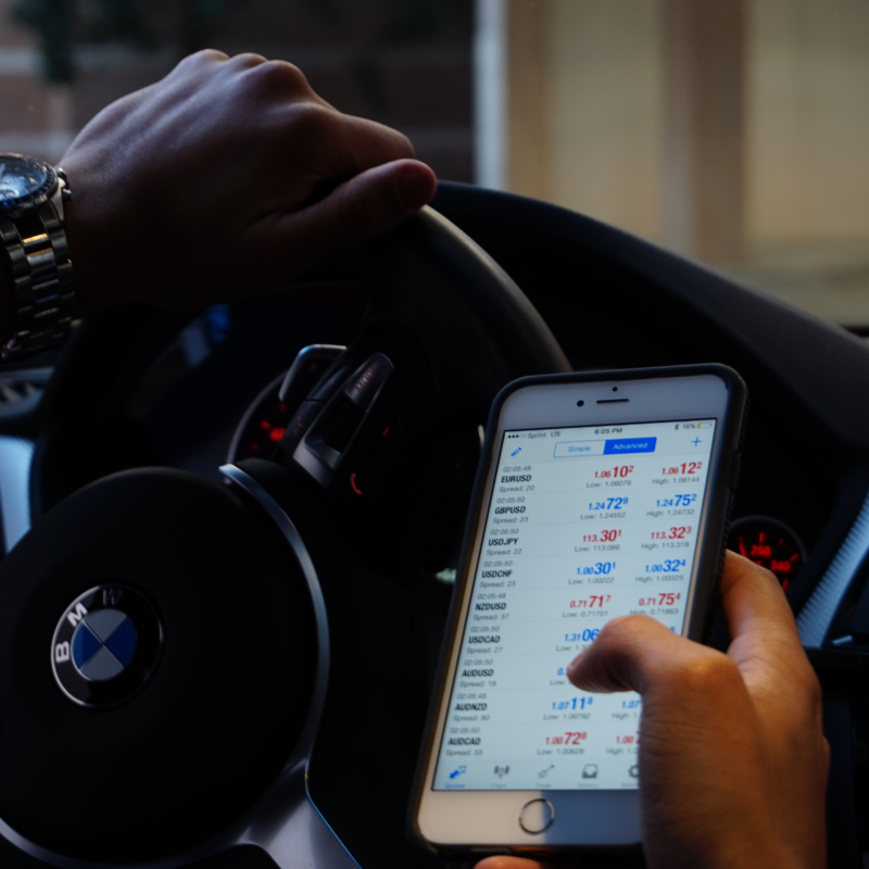 Driver in a BMW vehicle looking at a smartphone with financial information on screen, illustrating potential distracted driving.