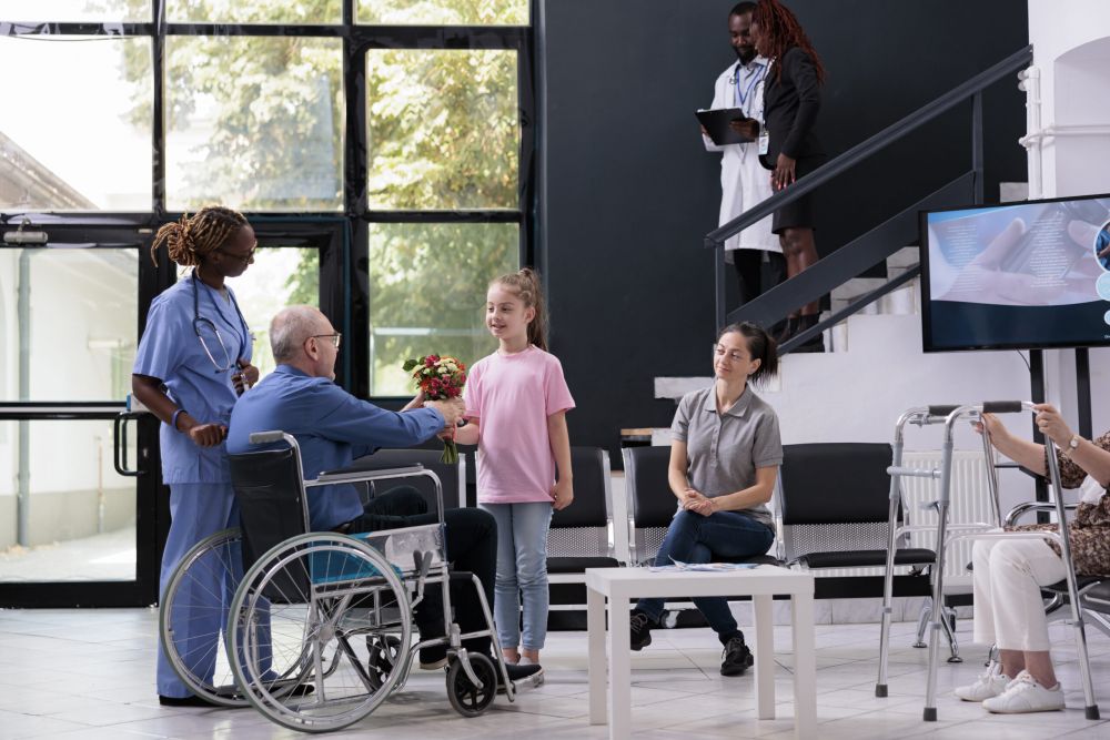 A young girl gives flowers to an elderly man in a wheelchair at a nursing home, while a nurse and family members look on with smiles.