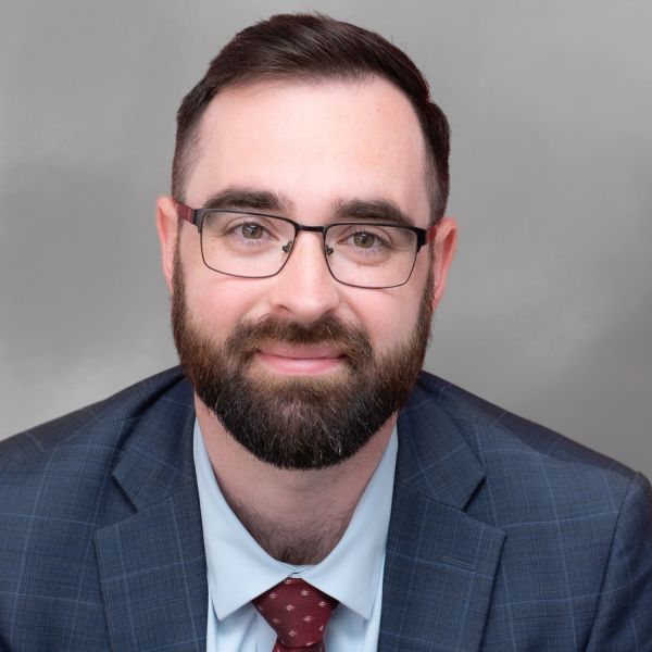 Hombre profesional con traje y gafas, con barba, sonriendo sobre un fondo gris.