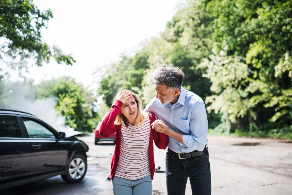 A distressed woman holding her head after a car accident, supported by a concerned man, with damaged vehicles in the background.