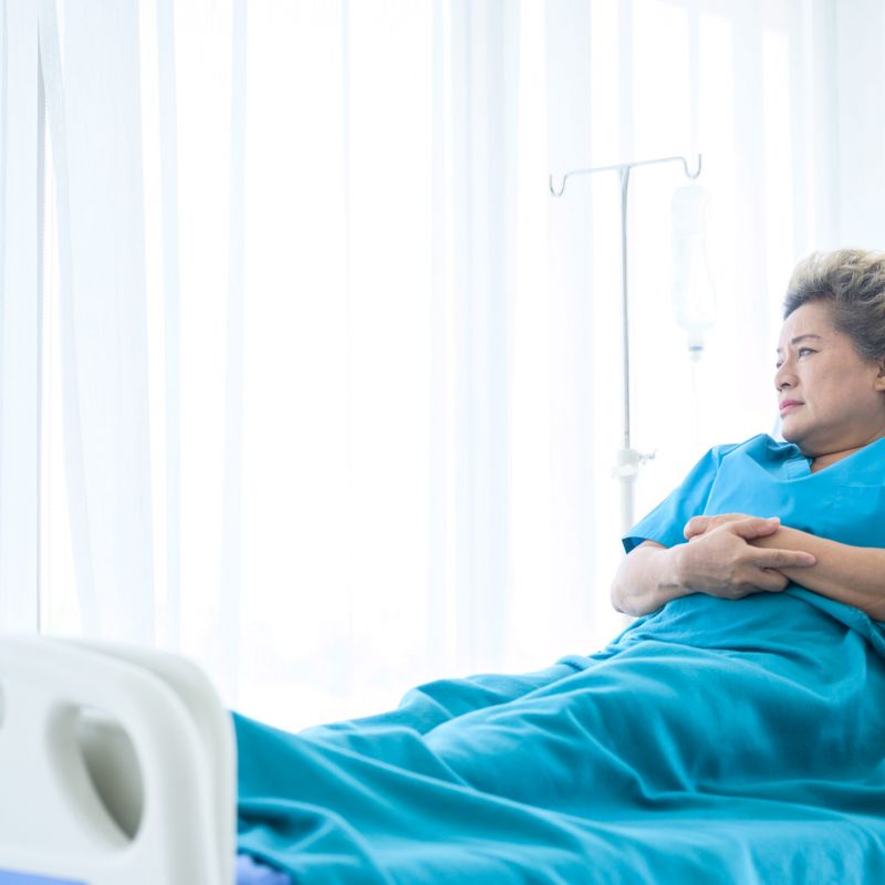 Elderly woman lying in a hospital bed, wearing a blue gown and gazing out the window, symbolizing nursing home neglect and isolation.