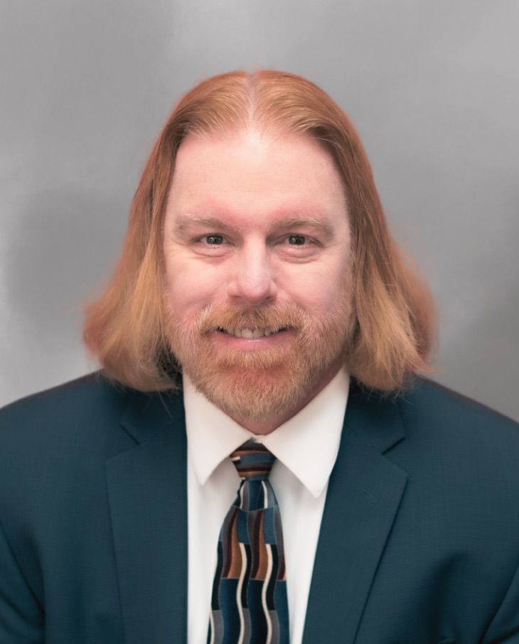 Jonathan DeLoatche with long hair and beard in suit and tie against gray background, smiling confidently.