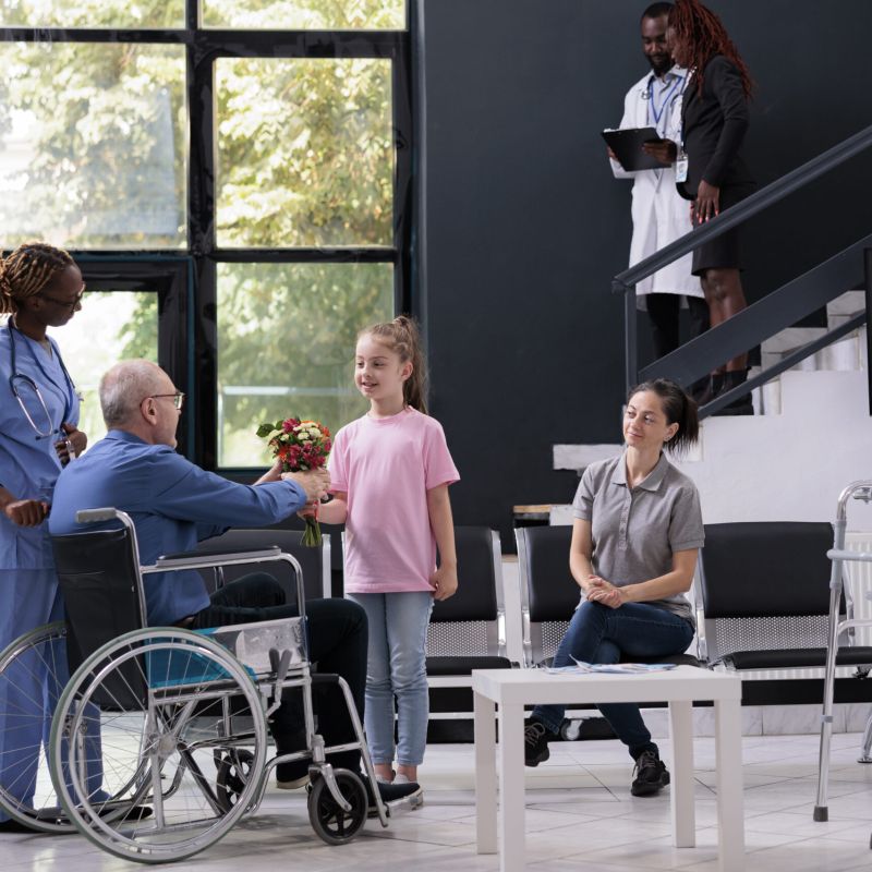A young girl gives flowers to an elderly man in a wheelchair at a nursing home, while a nurse and family members look on with smiles.