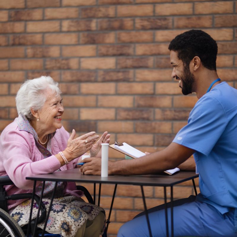 A smiling elderly woman in a wheelchair engaging in a friendly conversation with a healthcare worker in blue scrubs, discussing care plans.