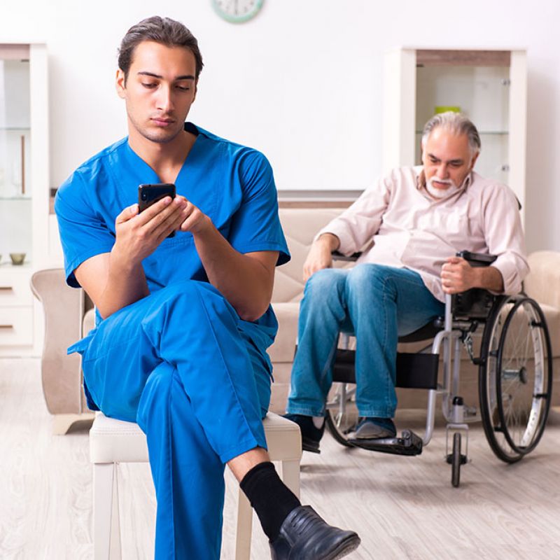 Caregiver in blue scrubs on phone while an elderly man in a wheelchair observes, in a home setting.