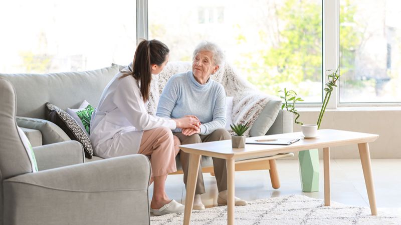 Caregiver and elderly woman sitting in a cozy living room, having a supportive conversation.