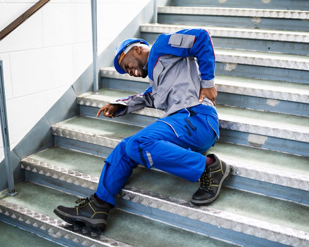 Worker in overalls and helmet falls on stairs, clutching back in pain. Workplace safety accident concept.