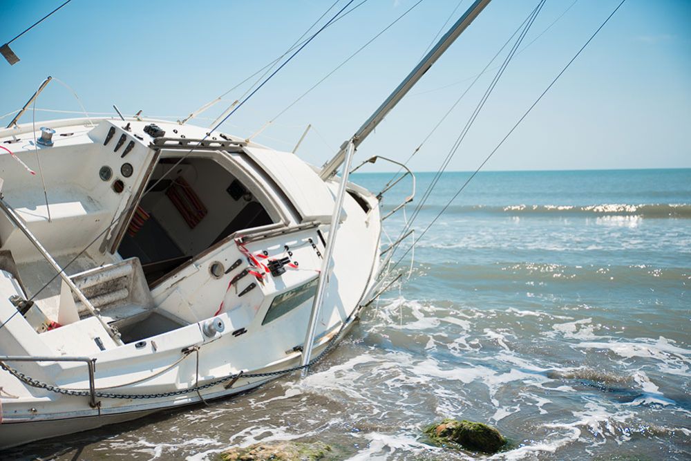 Sailing boat washed ashore, partially submerged in water under a clear blue sky at the beach.