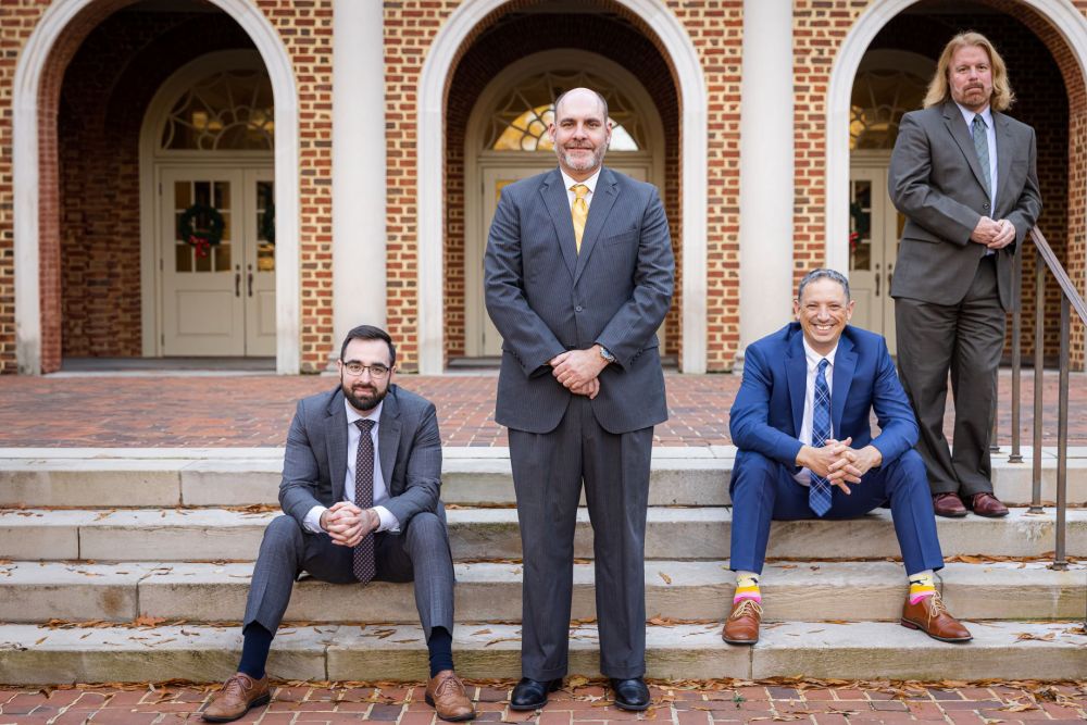 Four men in suits pose in front of a brick building with arches, two sitting and two standing on steps.