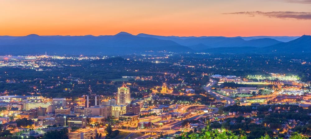 Cityscape of Roanoke, Virginia at sunset, with mountains in the distance and a vibrant skyline illuminated by city lights.