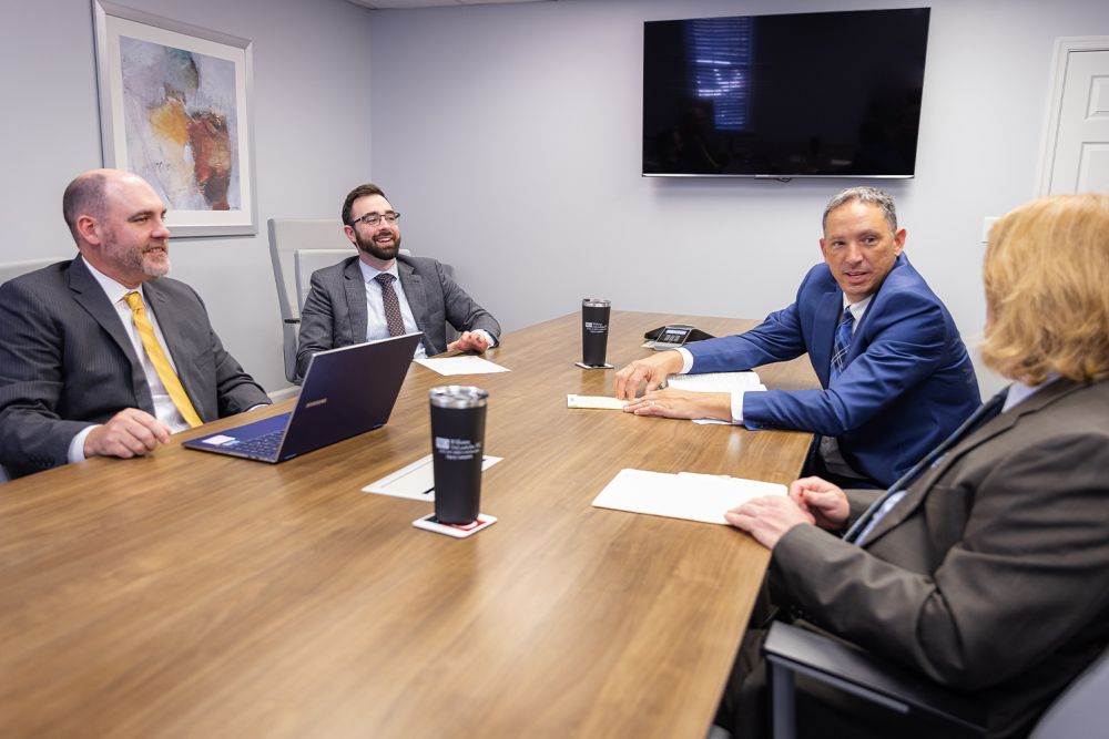 Four professionals in a business meeting room discussing around a table with laptops and documents.