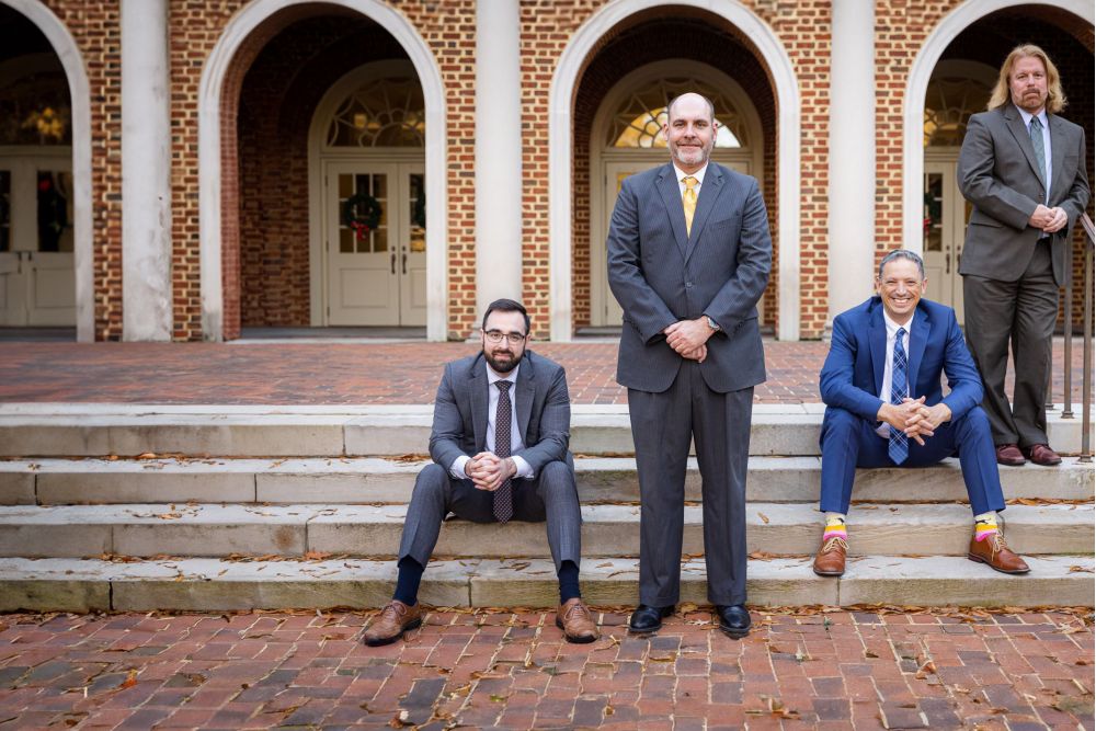 Four men in suits pose in front of a brick building with arches, two sitting and two standing on steps.