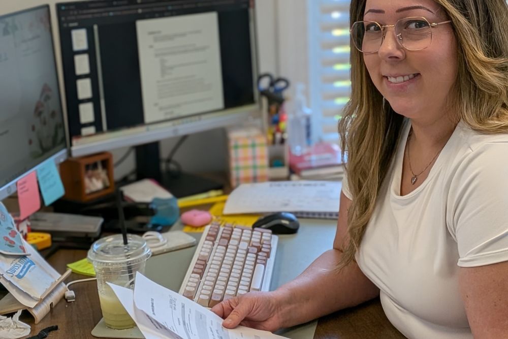 Woman at desk reviewing documents with dual monitors and office supplies.