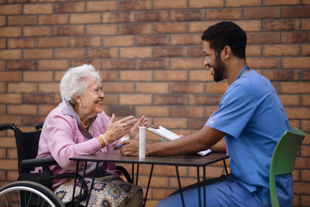 A smiling elderly woman in a wheelchair engaging in a friendly conversation with a healthcare worker in blue scrubs, discussing care plans.