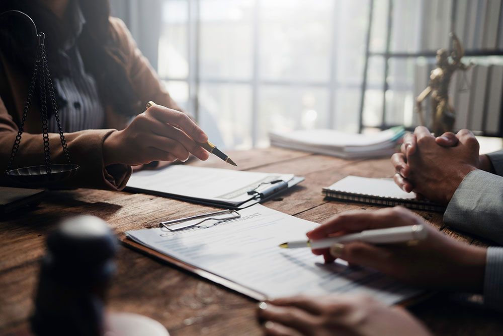 Business meeting with people discussing documents at a wooden table, with justice scales and statue in the background.
