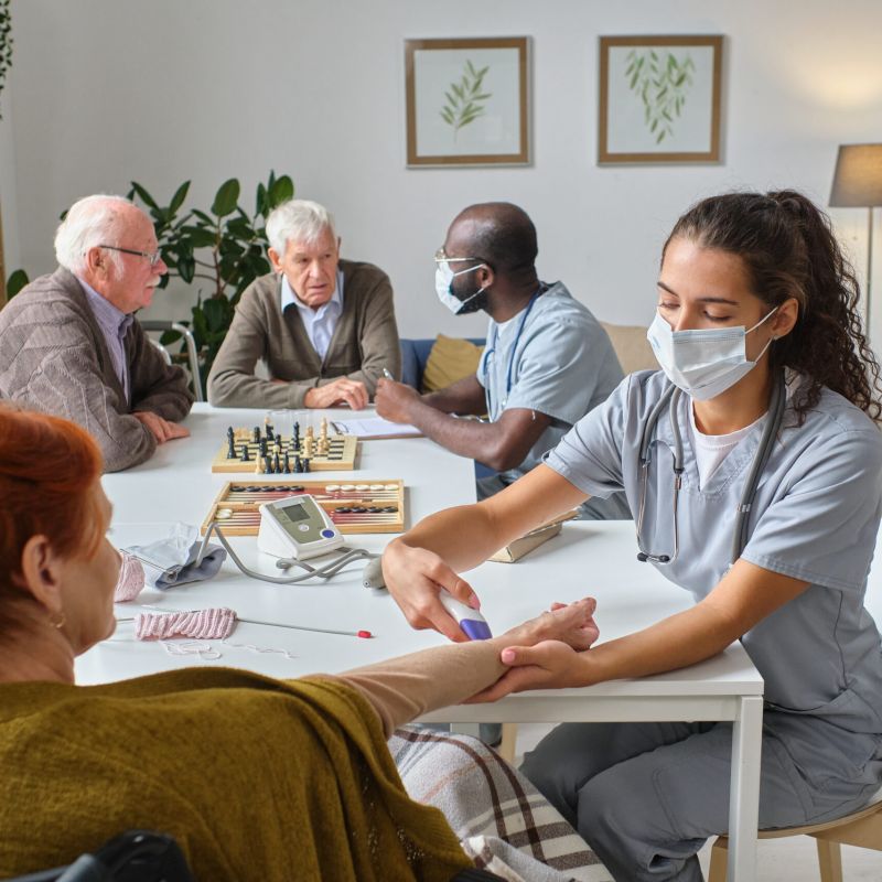 A nurse in scrubs checks a senior woman's blood pressure, while other elderly residents and staff interact at a nearby table in a nursing home.