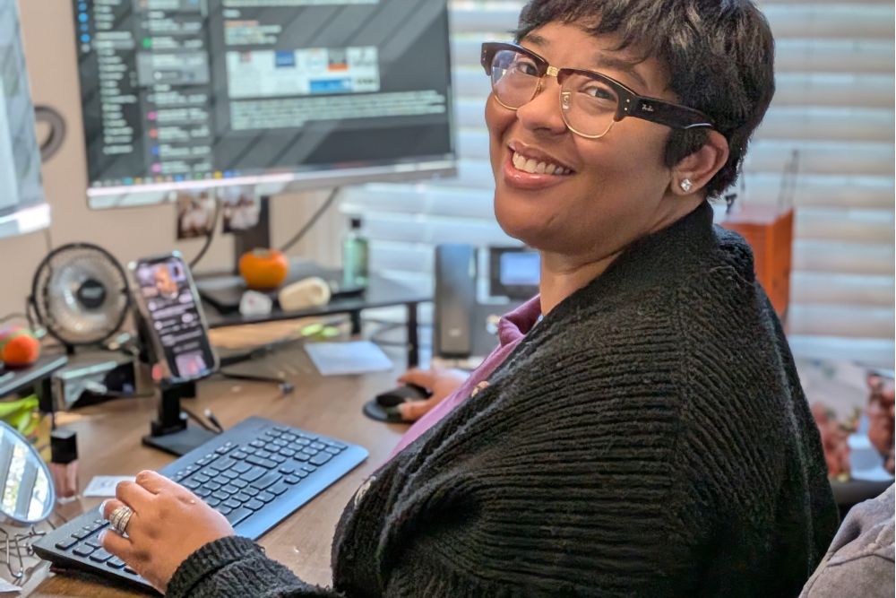 Smiling person using a computer in a home office with multiple screens and a cozy setup.