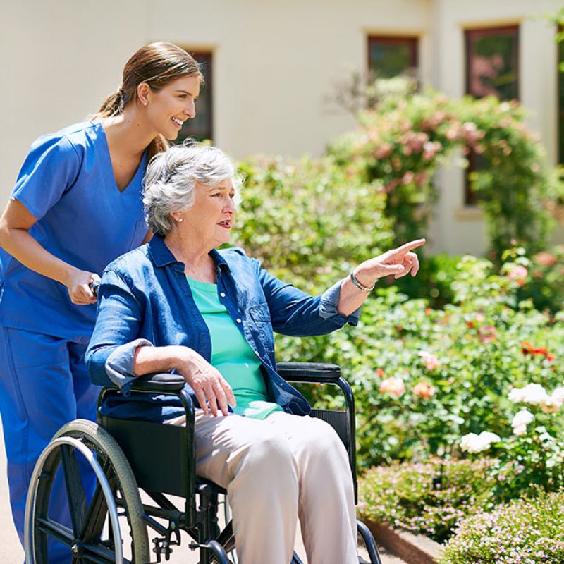 Caregiver assisting elderly woman in wheelchair through a garden, both smiling, surrounded by flowers and greenery.