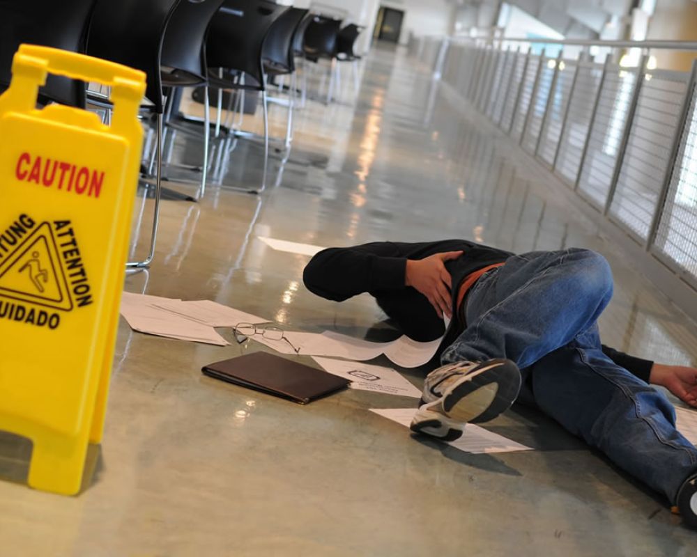 Person slipped on wet floor near caution sign in hallway, surrounded by scattered papers and belongings.