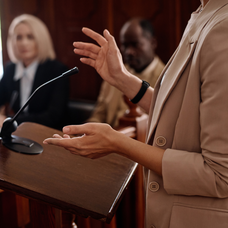Close-up of a woman gesturing while speaking at a courtroom podium, with two blurred figures, possibly legal professionals, in the background.