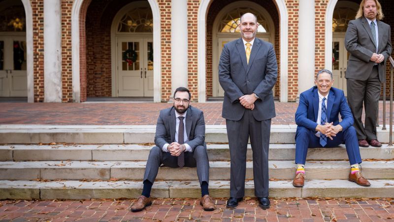 Four men in suits pose in front of a brick building with arches, two sitting and two standing on steps.