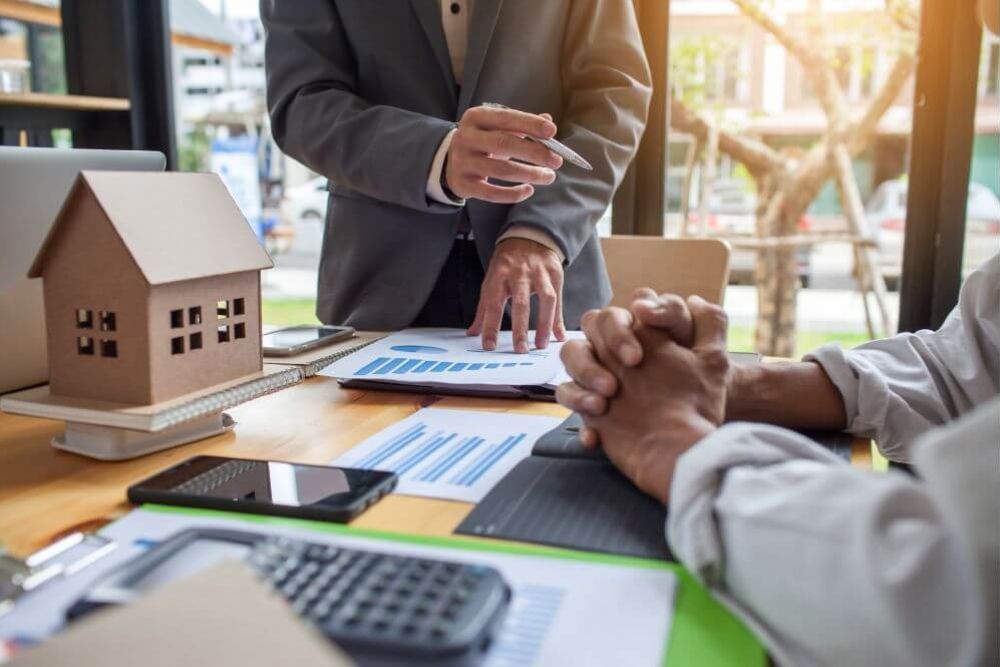 A person in a suit points at graphs on a table, while another person clasps their hands. Nearby, a small model house and various office tools are arranged on the desk.
